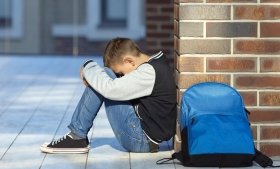 A child sits on the floor with their head down and back against a brick wall. A blue backpack sits next to them. Credit: Adobe Stock.