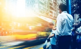 People stand in front of a street as a bus passes. Image credit: Adobe Stock