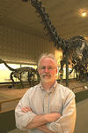 Portarit of Michael J. Donoghue. He stands, cross-armed, in front of huge dinosour skeletons from the main exhibition of the Peabody Museum. 