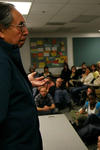 A native-american man stands and speaks in front of a classroom. He wears rounded glasses and extends a hand, palm facing up, to the crowd. 
