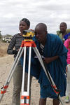 Six people stand doind different activities. One of them looks through the eye-piece of equipment mounted on a tripod. Behind them, clear skies can be seen.