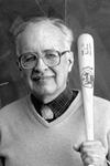 Robert K. Adair stands in front of a blackboard holding a baseball bat. A diagram of a baseball and bat are drawn in chalk on the blackboard.