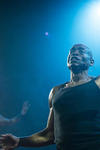 A man stands under stage lighting. Only his torso can be seem. Mid-word and closed-eyed, he lifts his muscular left arm and opens his hand, palms facing forward. The whole picture has a blue tone. In the distance, a picture of the same man yet from a different angle appears. 
