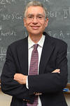 Yoram Alhassid in a black suit and patterned tie, standing, smiling, arms folded, in front of a chalkboard with mathematical symbols written